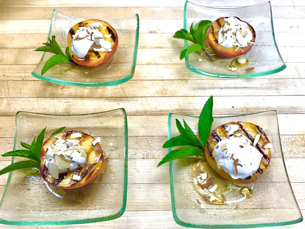 Grilled peaches topped with ice cream, almond slivers, and mint, arranged on glass plates.