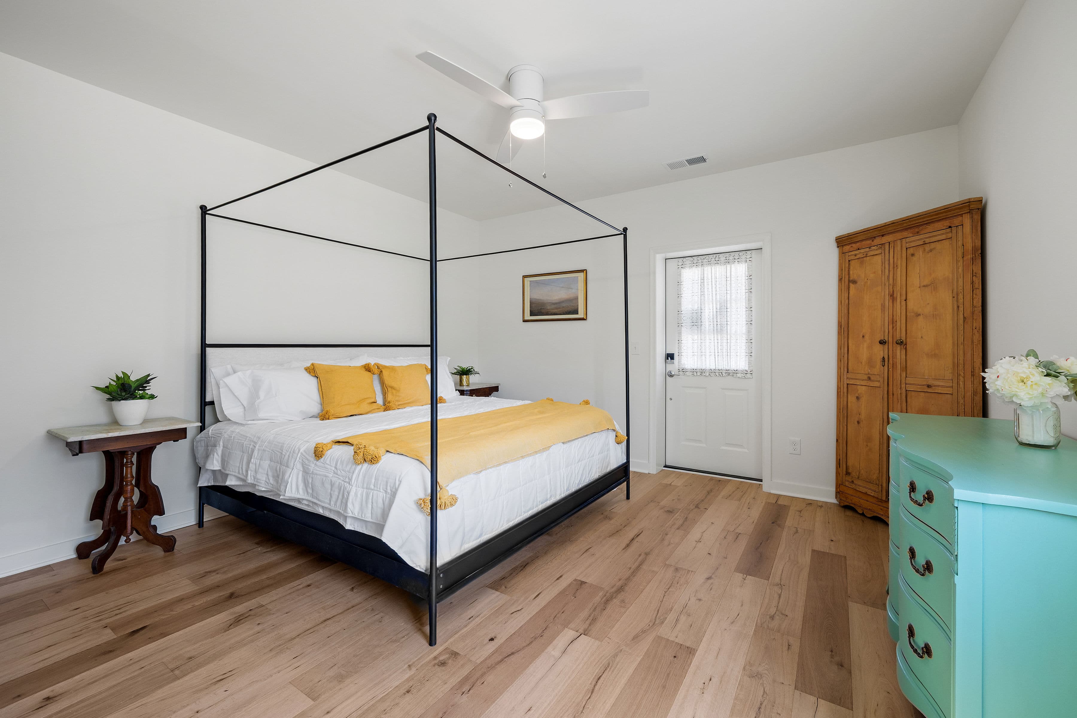 A bright, minimalist bedroom featuring a four-poster bed with yellow accents, a wooden wardrobe, and a cheerful blue dresser.