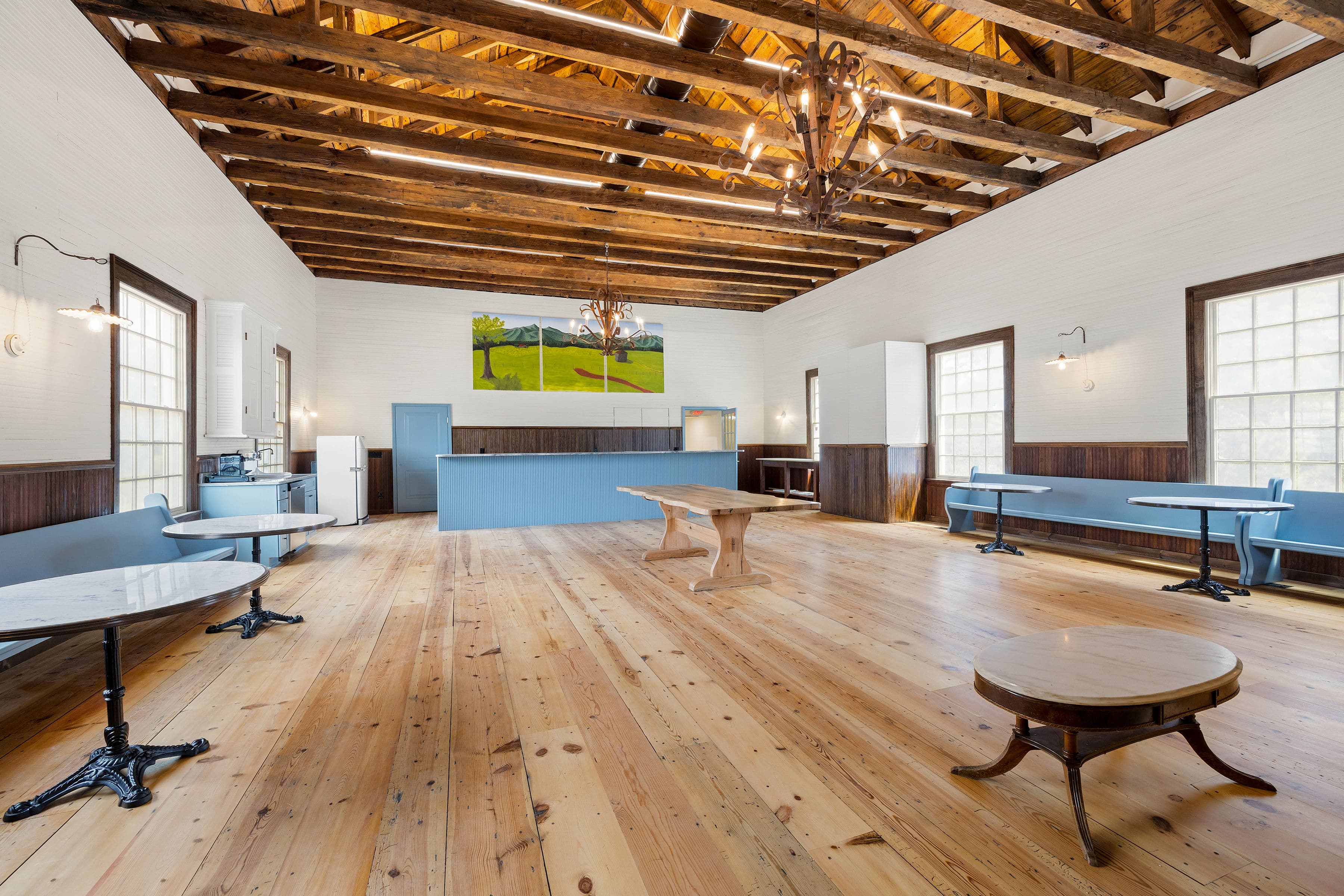 A spacious, well-lit room with wooden floors, high ceilings, and a blue bar counter, furnished with several round tables.