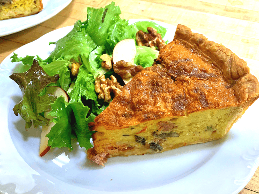 A slice of quiche served with a side of mixed greens, walnuts, and apple slices.