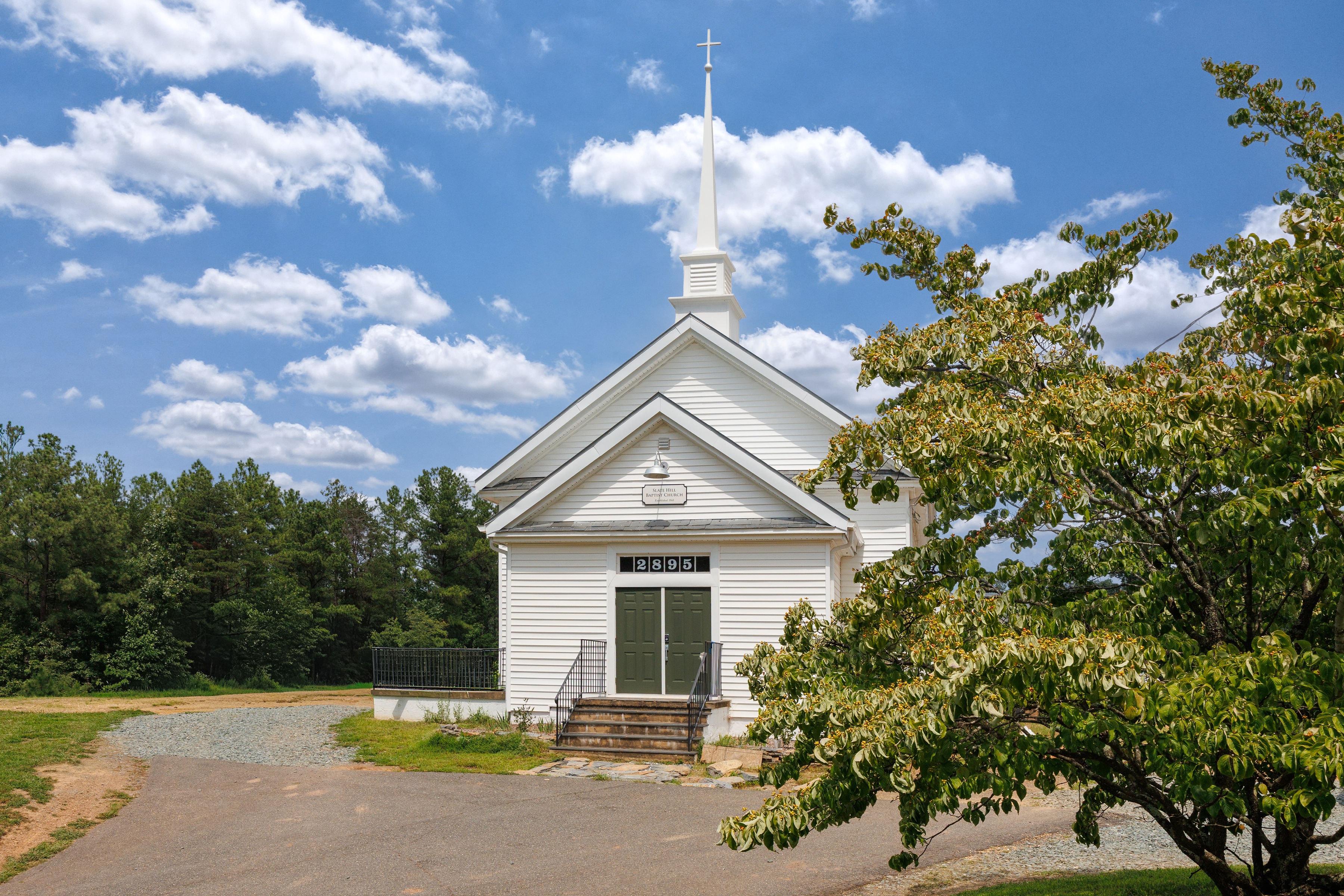 A white wooden church with a spire, surrounded by greenery under a blue sky with clouds.