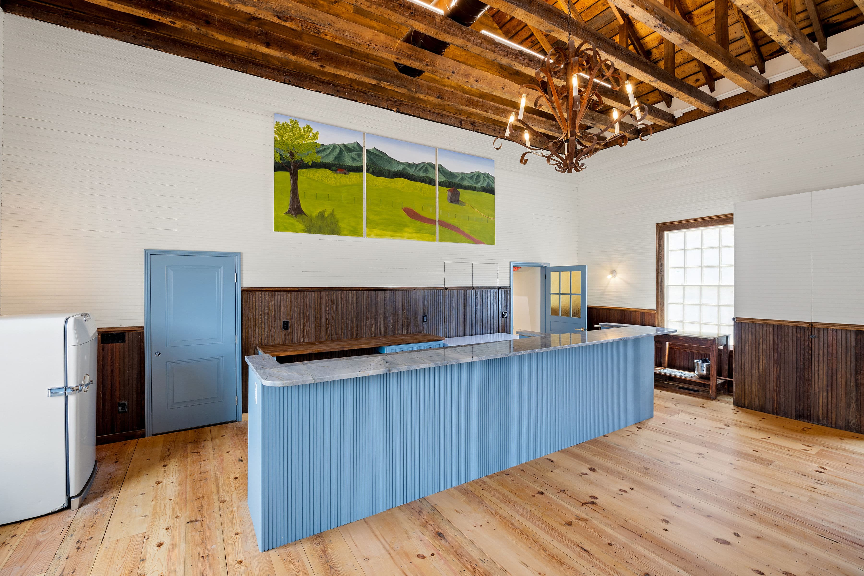 Bright interior featuring a blue bar counter, wooden beams, and landscape artwork on the wall.