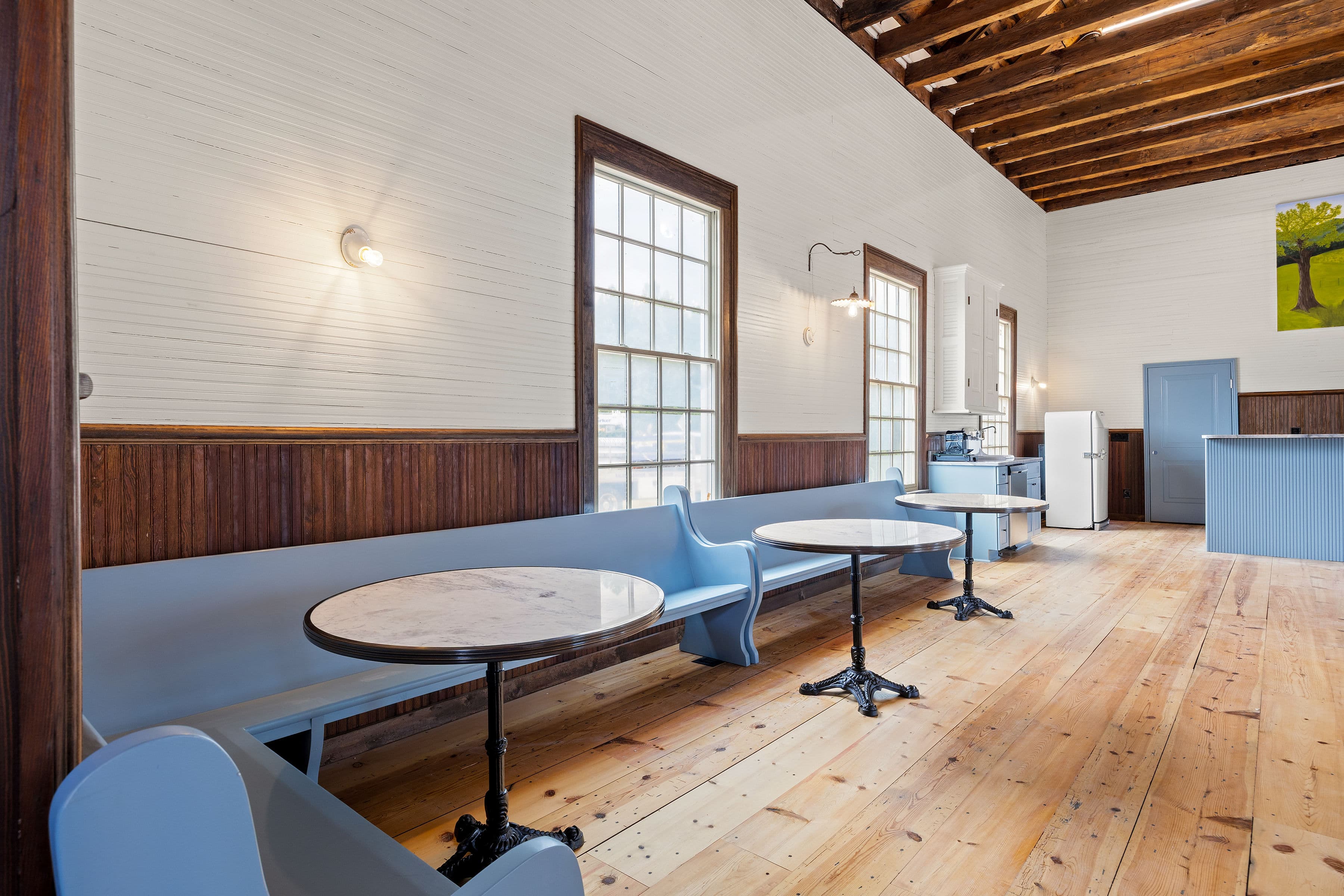 A spacious, well-lit café with wooden floors, blue seating, and natural light from large windows.