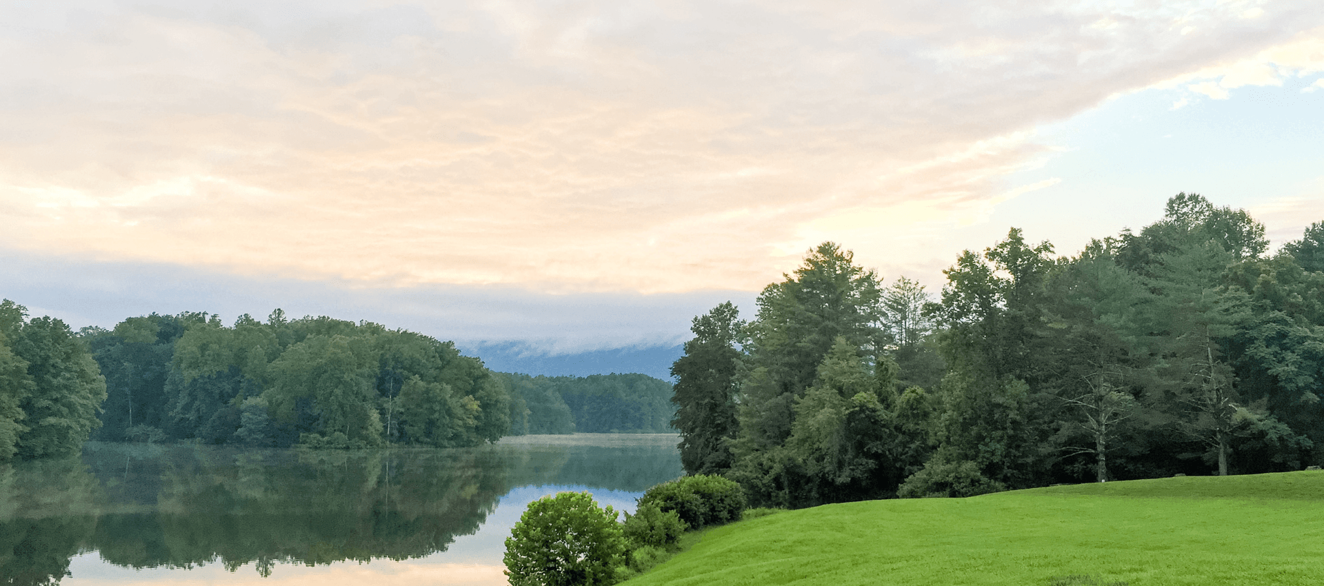Serene landscape featuring a calm lake surrounded by lush greenery and soft clouds in the sky.