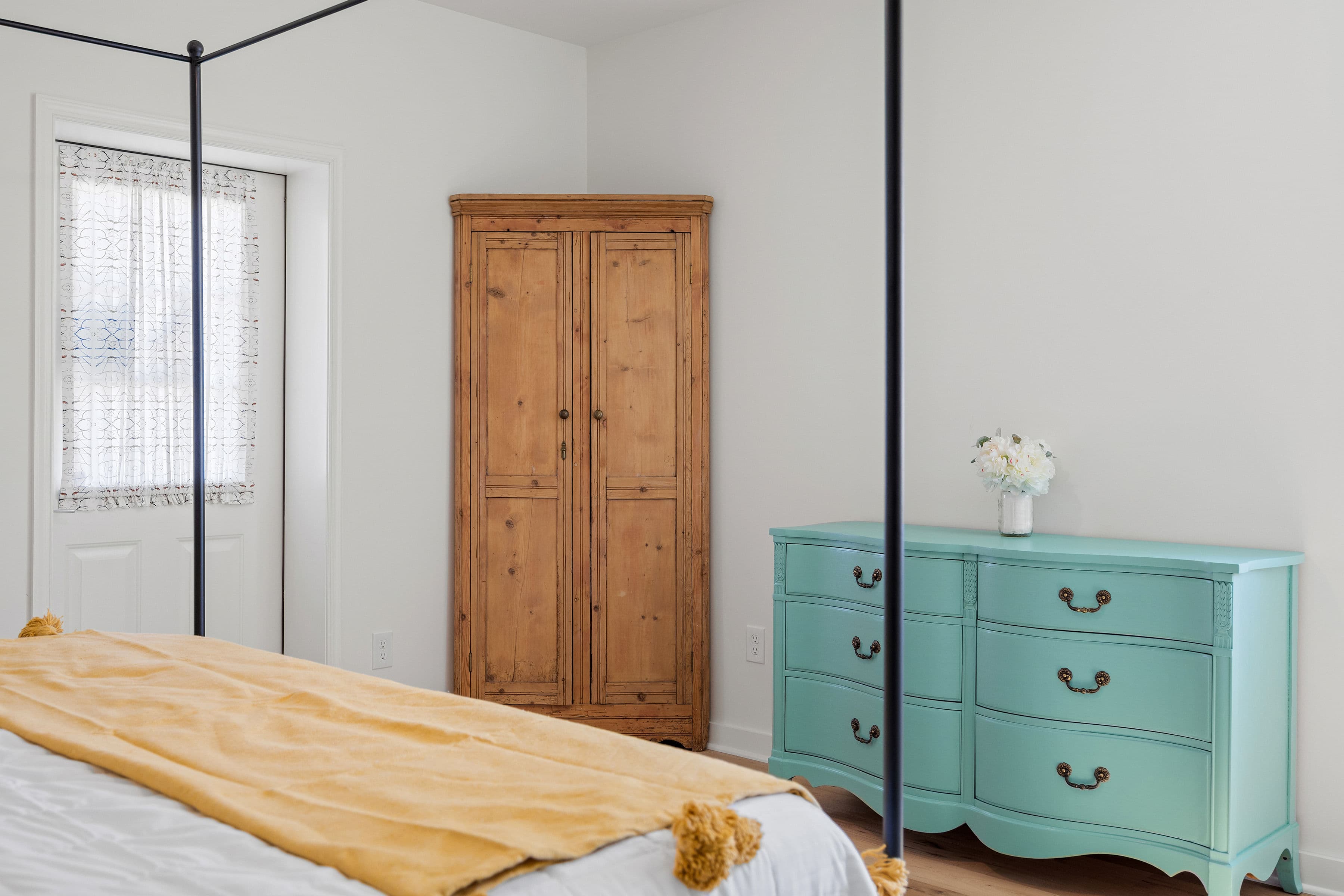 A cozy bedroom featuring a four-poster bed, wooden wardrobe, and a teal dresser with a vase of flowers.