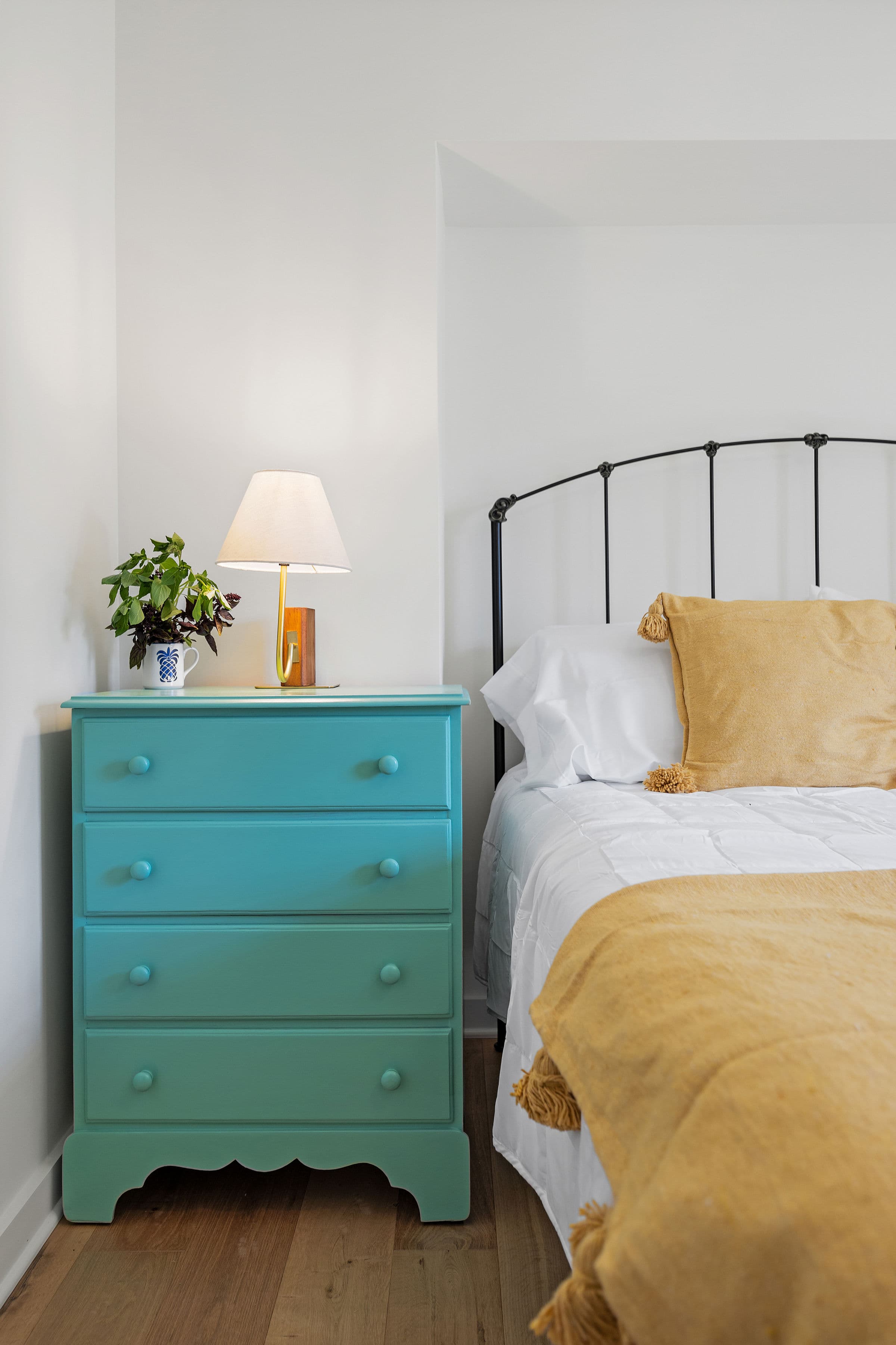 A turquoise bedside dresser with a lamp and a plant next to a neatly made bed with yellow accents.