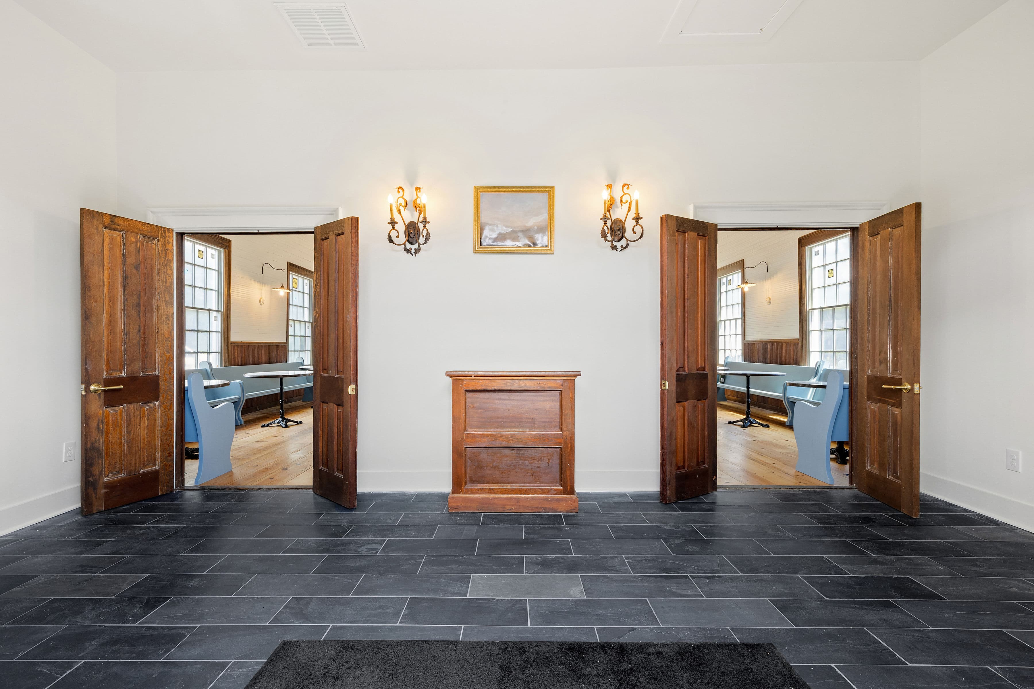 A spacious foyer with two open wooden doors leading to rooms, featuring a central wooden cabinet and wall sconces.
