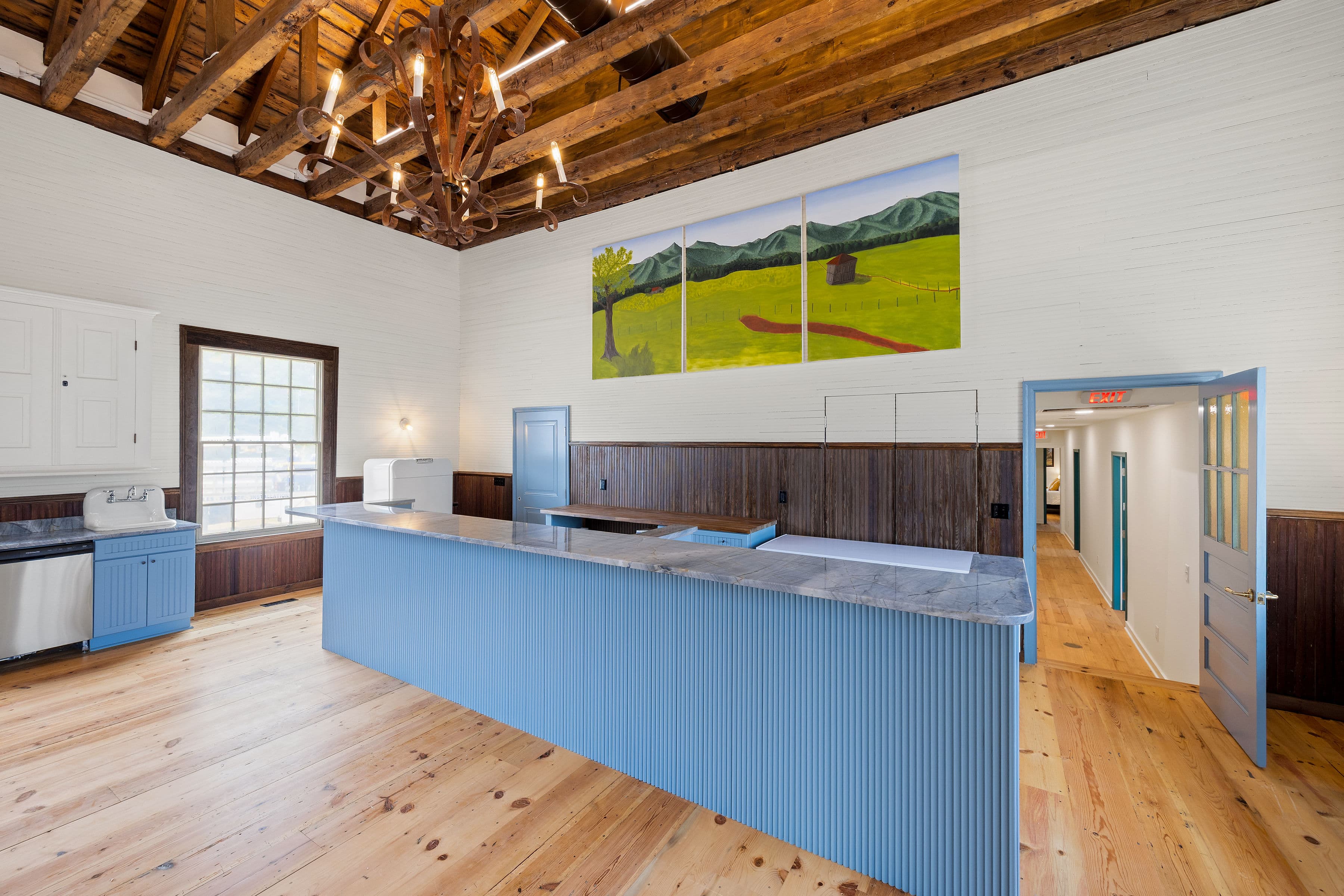 Modern kitchen with a blue island, wooden beams, and large windows showcasing a mural of mountains and trees.