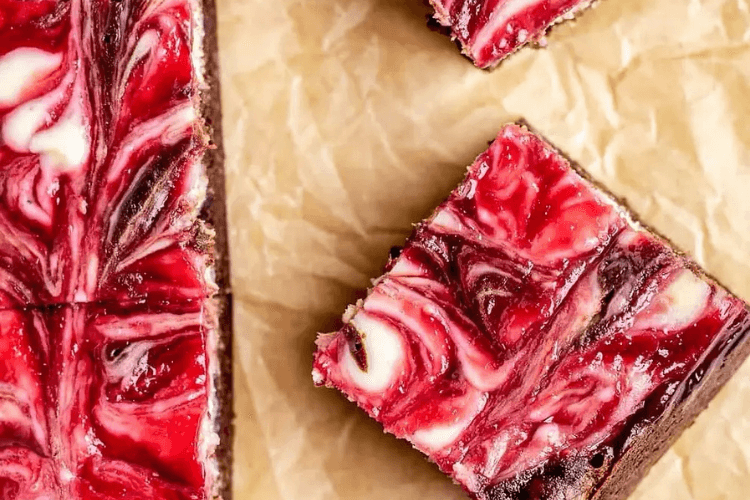 A close-up of red and white swirled cheesecake brownies on parchment paper.