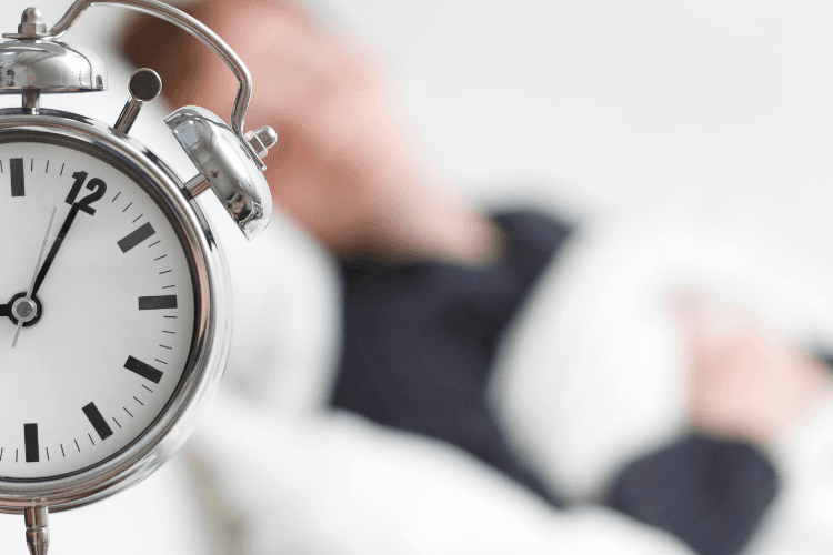 A silver alarm clock is in focus with a blurred person sleeping in the background.