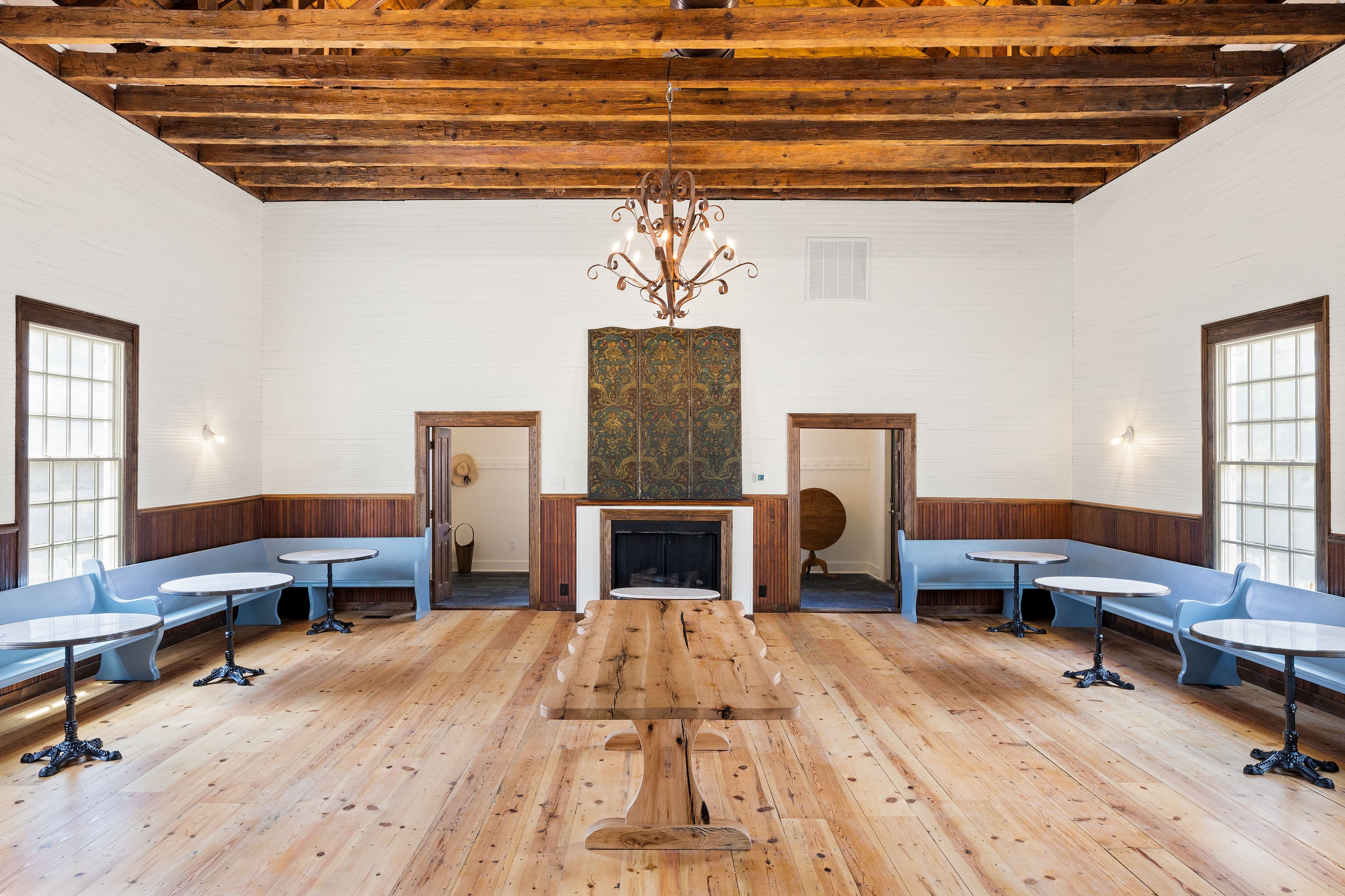 A spacious room with wooden floors, a long table, circular tables, and a chandelier, featuring blue seating along the walls.
