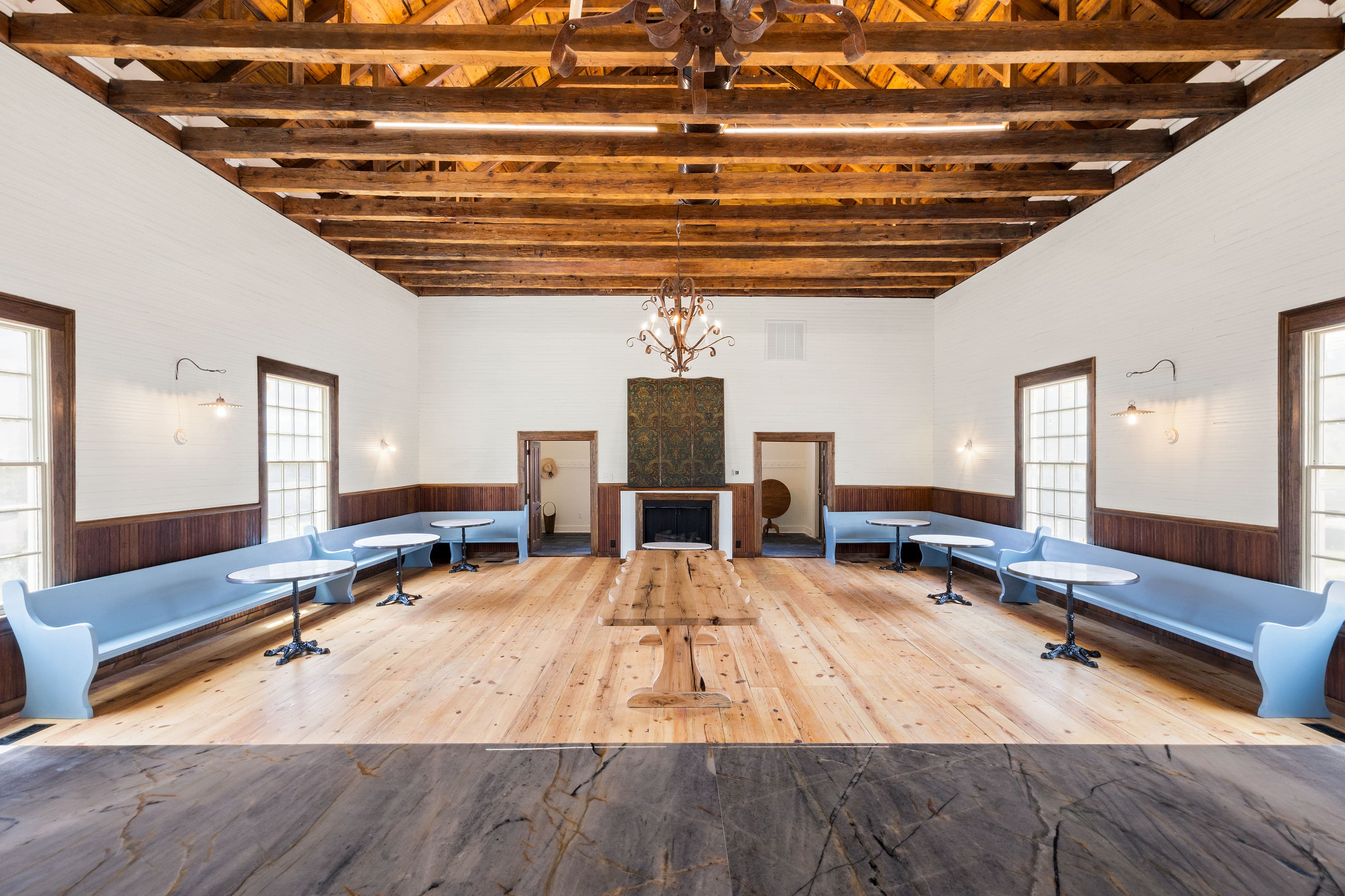 A spacious, brightly lit room with wooden beams, featuring blue seating and tables arranged around a central wooden table.
