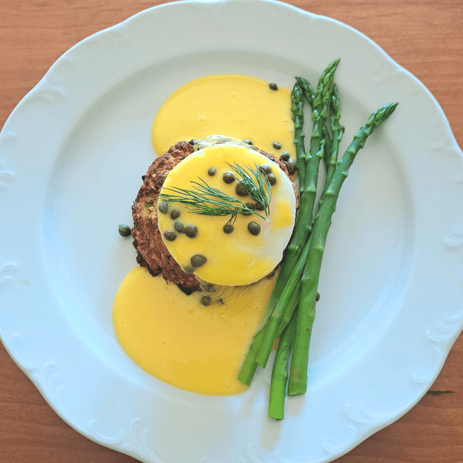 A plate featuring a poached egg on a savory base, topped with hollandaise sauce and garnished with capers and dill, alongside asparagus spears.