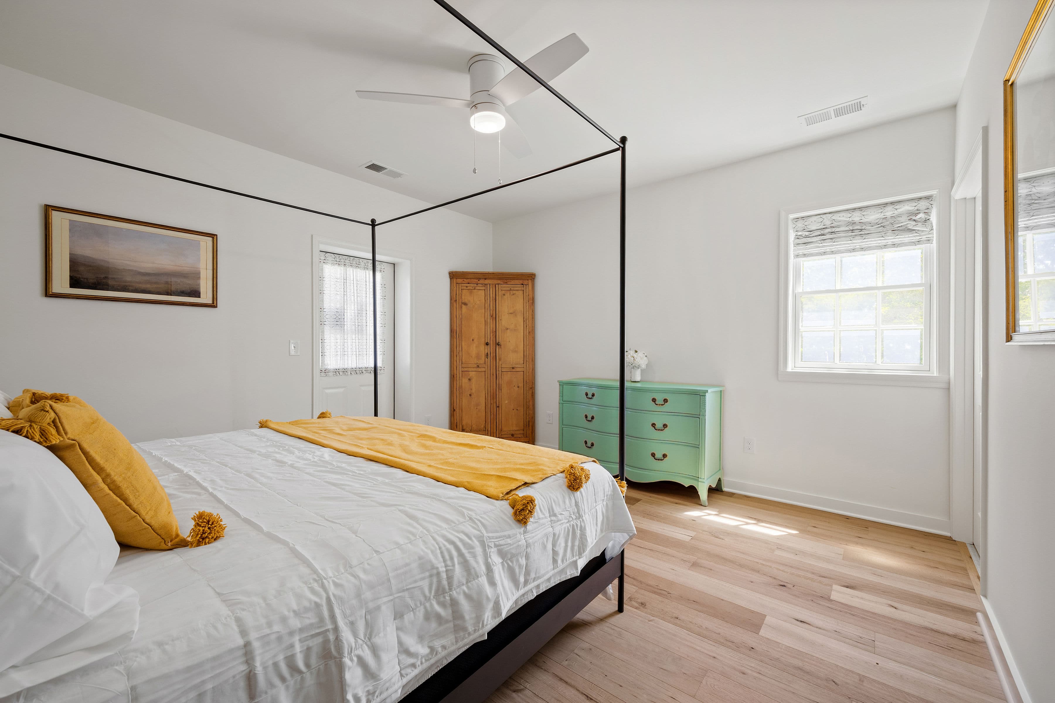 A bright and airy bedroom featuring a four-poster bed with a yellow throw, a green dresser, and large windows.