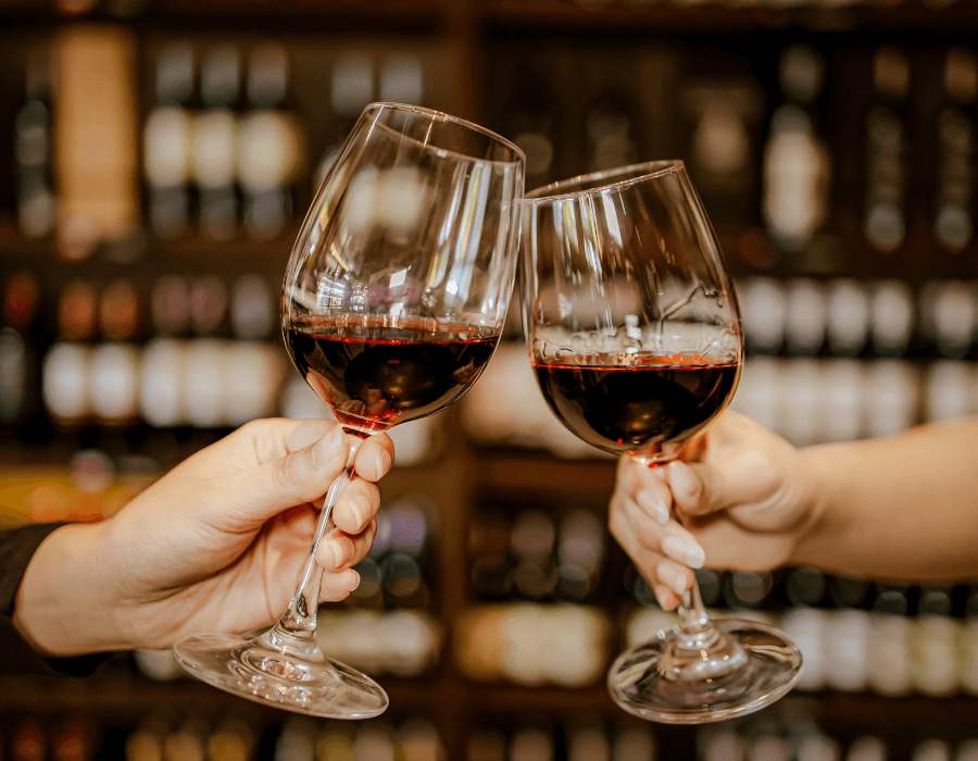 Two hands clink wine glasses filled with red wine against a blurred background of wine bottles.
