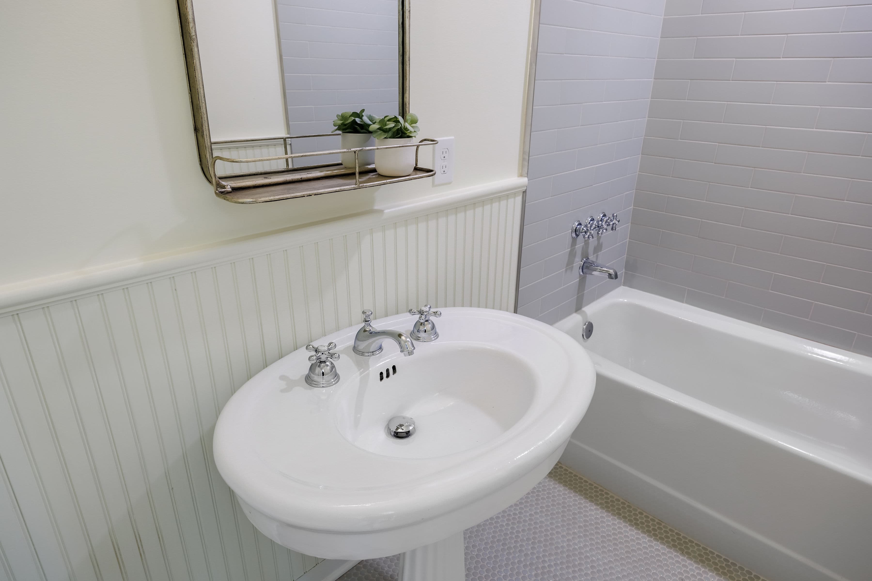 A white pedestal sink and tub in a clean, modern bathroom with a mirror and small plant.