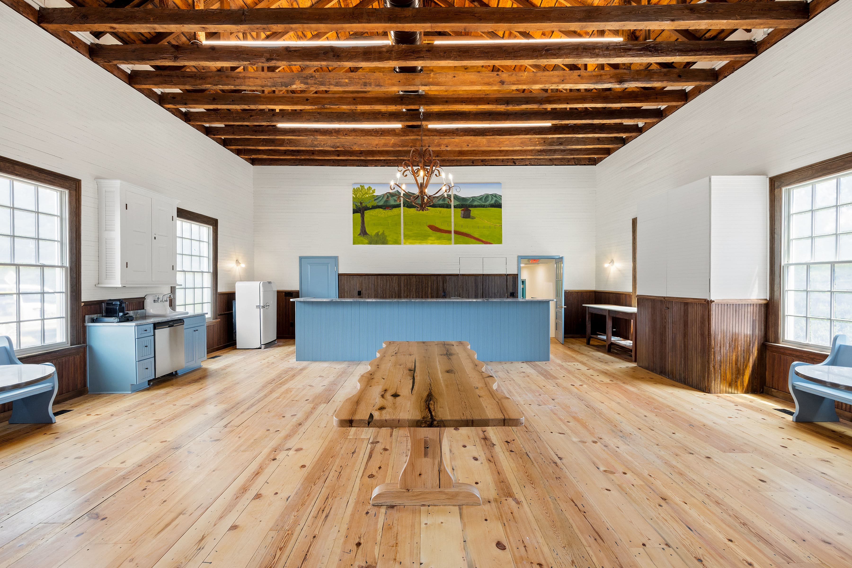 Spacious, rustic interior with wood beams, a long wooden table, and blue kitchen cabinetry.
